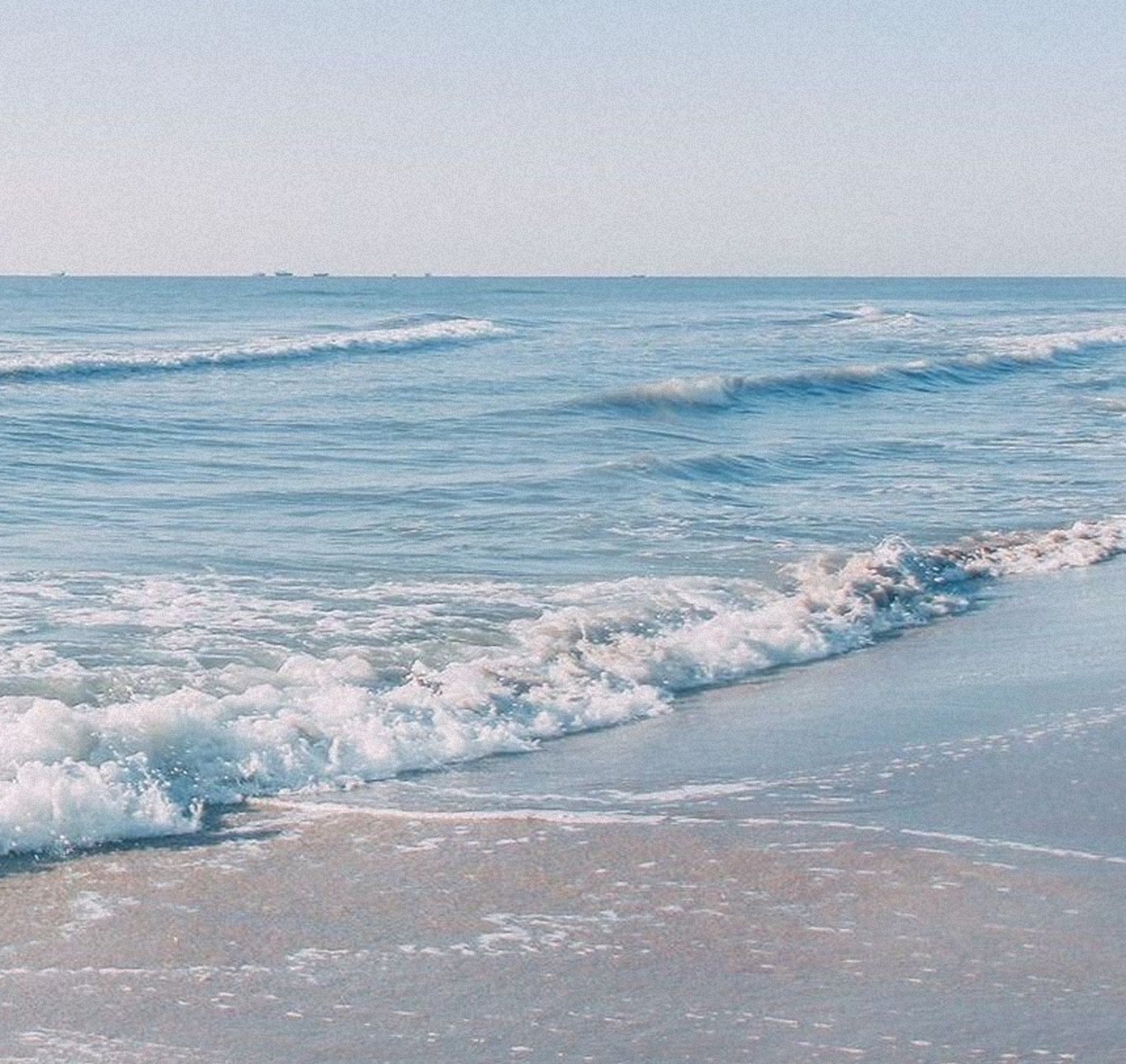 Gentle waves roll onto a sandy beach under a clear, pale blue sky. The horizon is visible in the distance, with calm sea stretching outwards and small whitecaps forming near the shore.