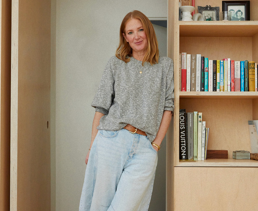 A woman with long red hair, wearing a gray sweater and light blue jeans, stands smiling next to a wooden bookshelf filled with books and framed photos.