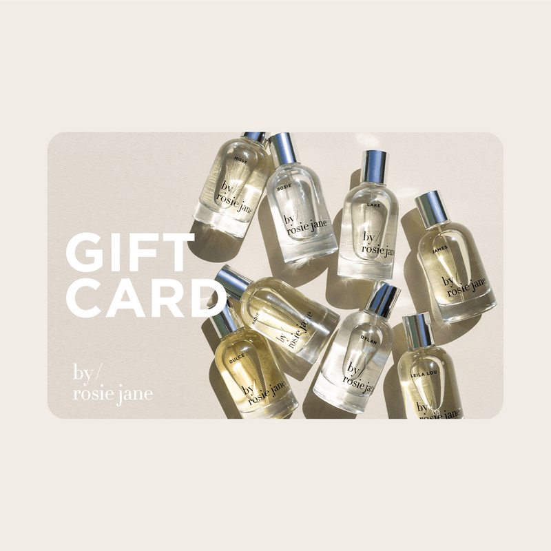 Several bottles of by/rosie jane perfumes are arranged on a light surface with sunlight casting shadows. The words FRAGRANCE DISCOVERY SET + $80 E-GIFT CARD and by/rosie jane appear on the left in white text, perfect for gifting.