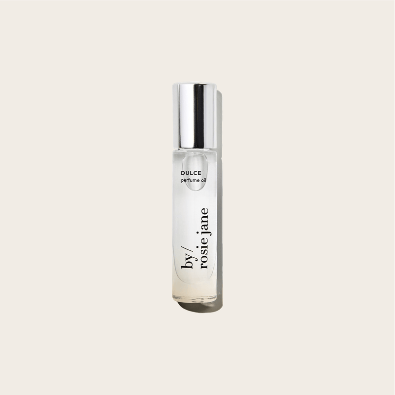 A clear glass byrosiejane DULCE PERFUME OIL bottle with a silver cap, featuring vanilla and nude musk notes, sits on a plain light beige background.