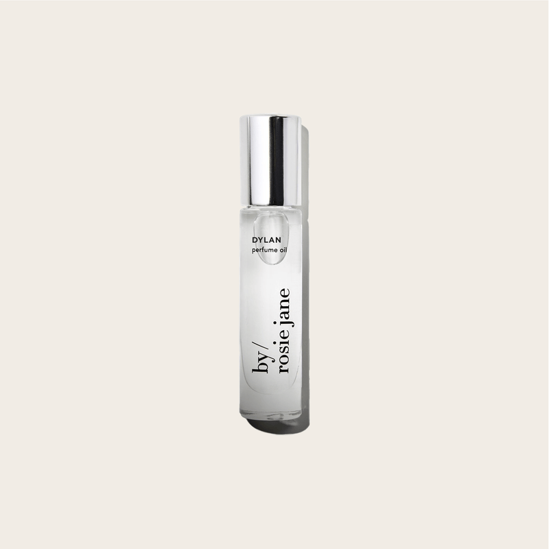 A clear glass bottle of byrosiejane DYLAN PERFUME OIL with a silver cap sits against a light beige background, featuring a vertical black label and evoking notes of white musk and cedar for a sense of sexual freedom.