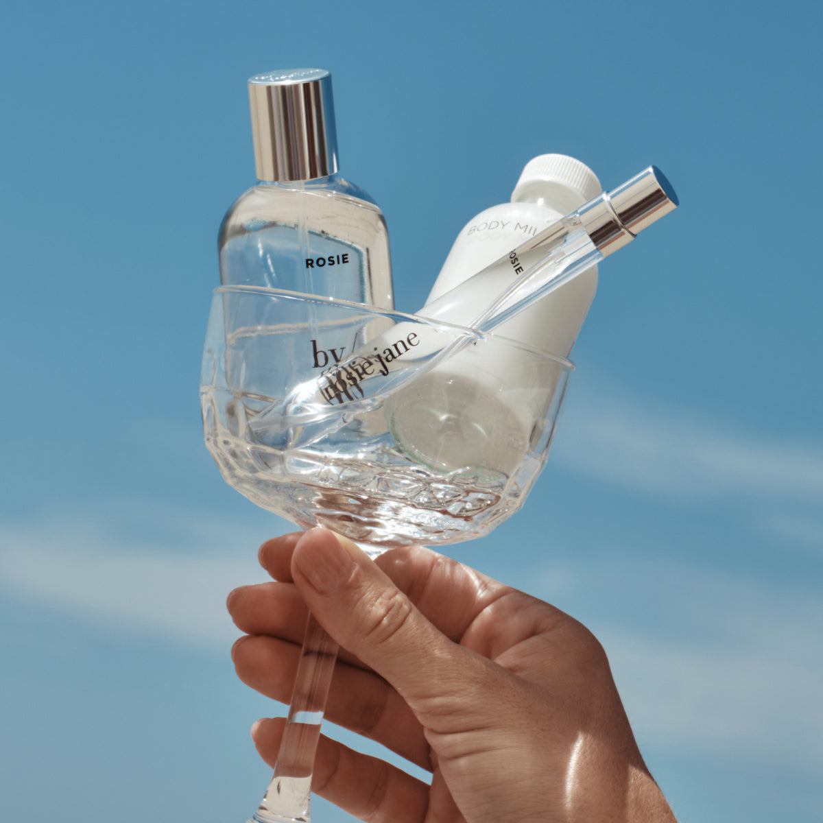A hand holds a clear stemmed glass containing two perfume bottles from by appearine, including a clear bottle labeled ROSIE and a white bottle, with a silver spray atomizer resting inside, against a bright blue sky background.