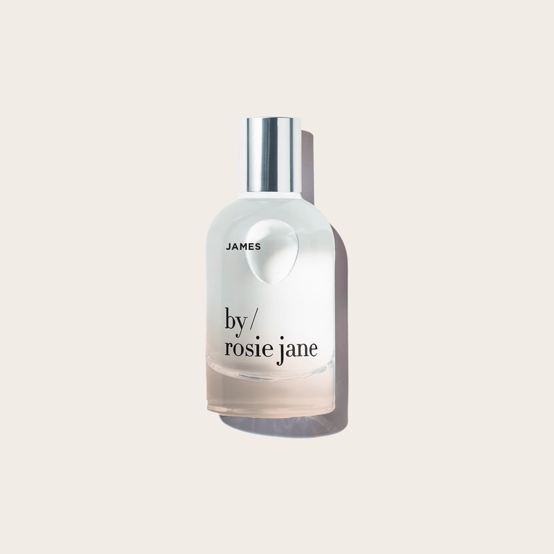 A clear glass JAMES PERFUME bottle by byrosiejane, featuring a silver cap and label, rests on a light beige background. Delicate gardenia notes linger as the bottle casts a soft shadow to the right.