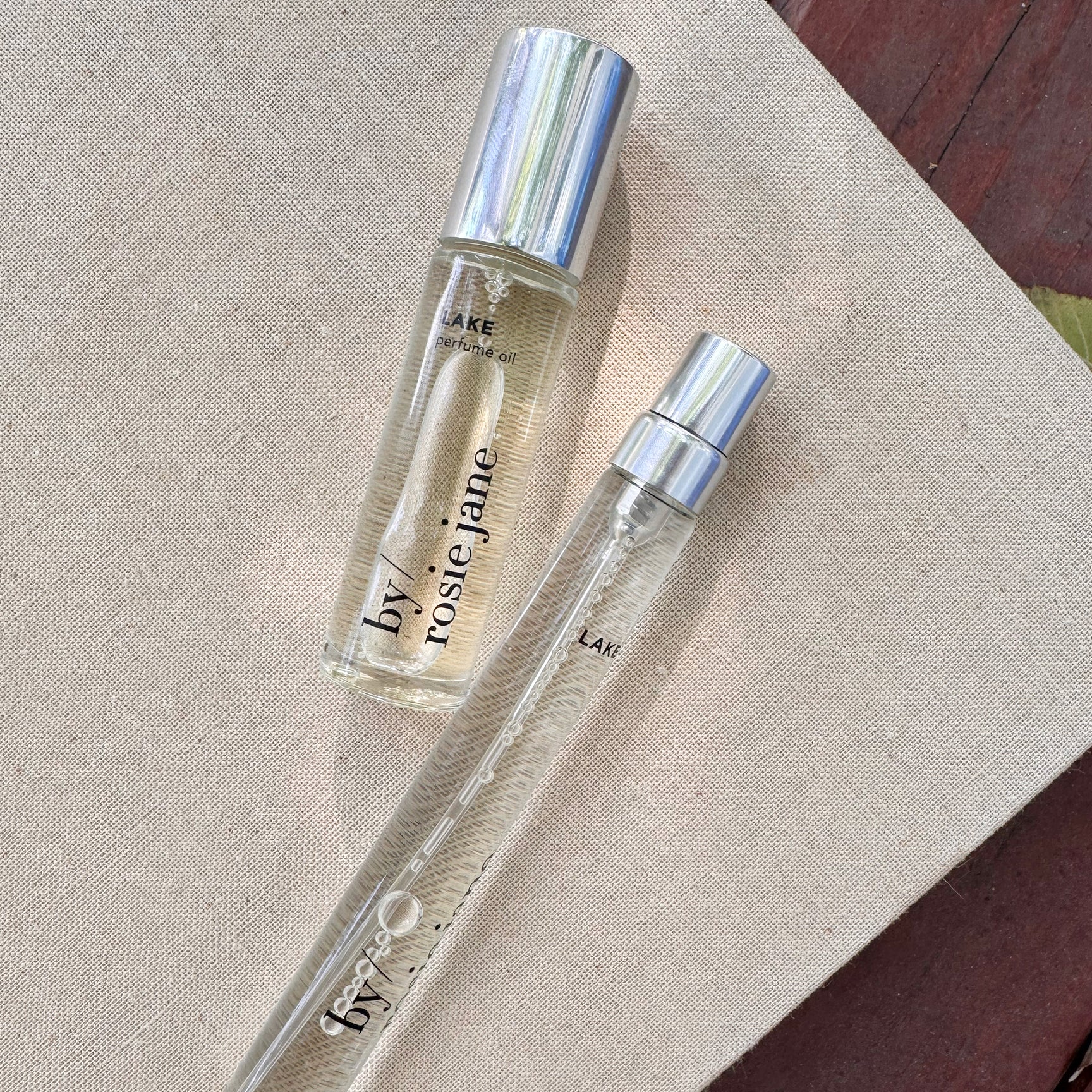 The LAKE 2 WAYS TRAVEL SET by byrosiejane, featuring two clear glass bottles with silver caps and a Lake Tahoe inspired scent, is displayed on a beige textured surface against a wooden background.