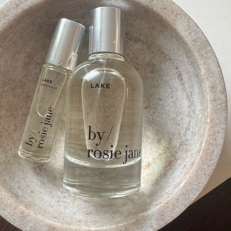 The LAKE LAST ALL DAY SET by byrosiejane features two clear glass bottles—a small Perfume Oil and a larger Eau de Parfum—both with shiny silver caps, nestled in a round stone dish for lasting fragrance.
