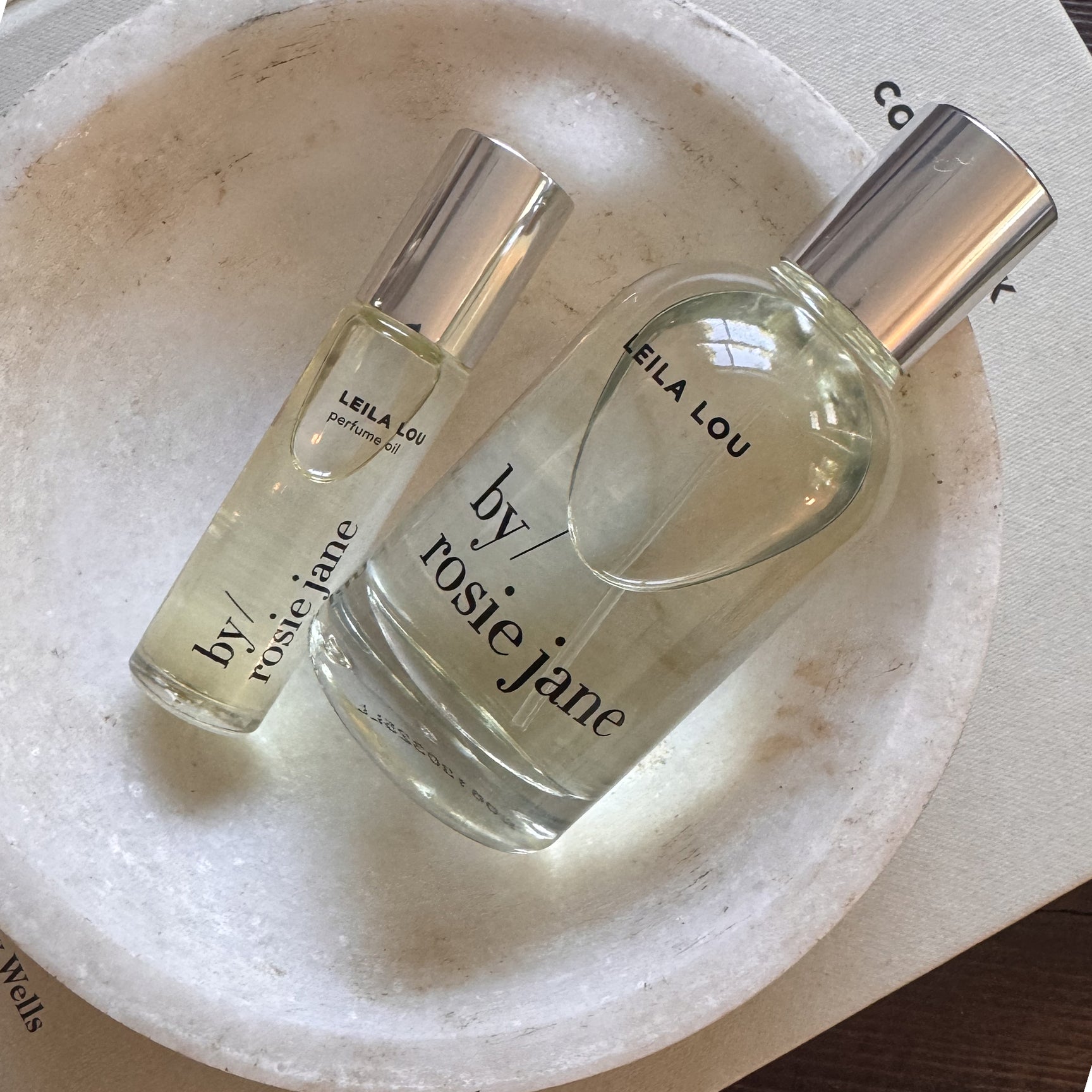 The LEILA LOU LAST ALL DAY SET by byrosiejane, featuring two clear glass bottles, sits on a round white marble dish atop a light-colored book.