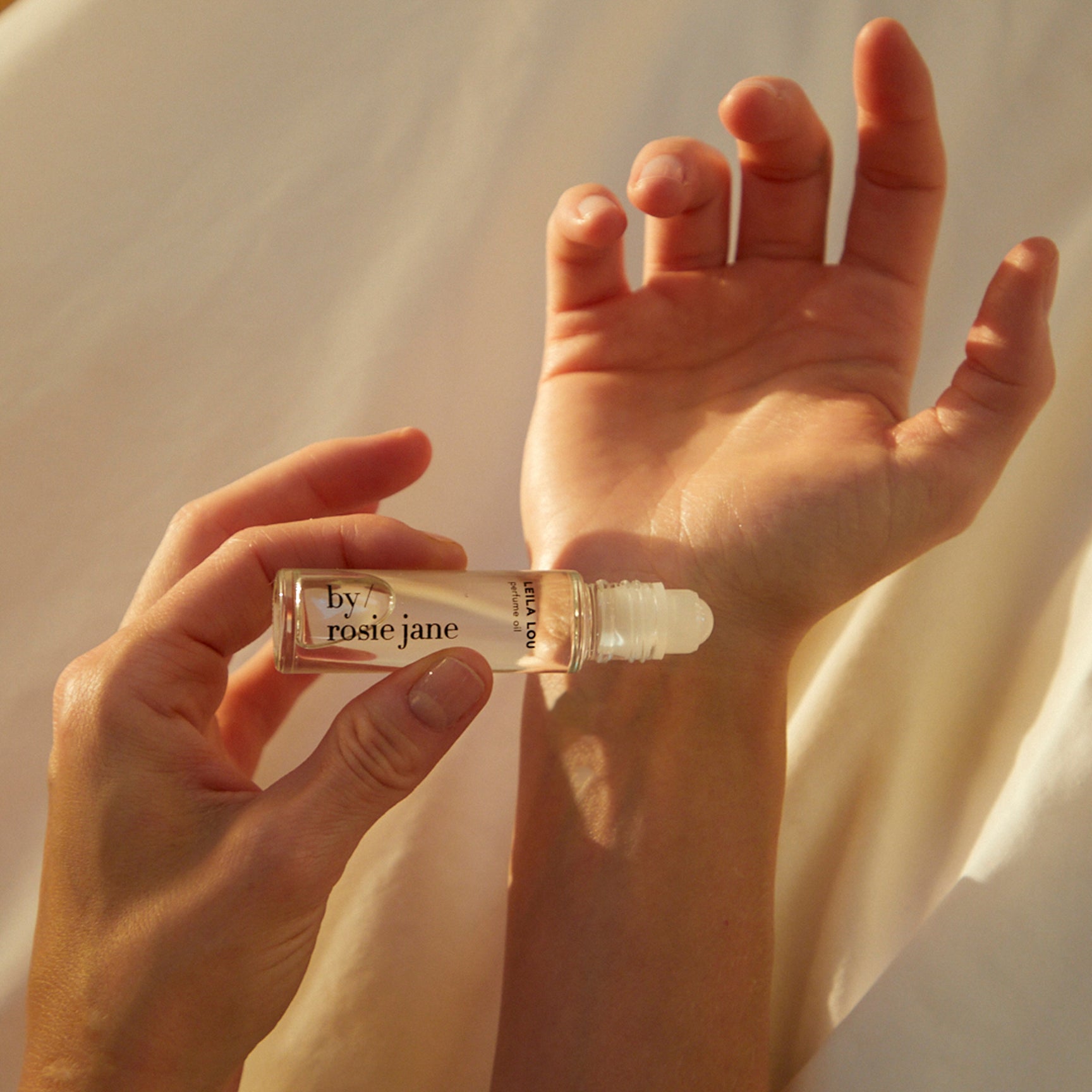 A hand rolls byrosiejanes LEILA LOU PERFUME OIL onto anothers wrist, releasing a delicate jasmine fragrance as soft natural light illuminates the scene.