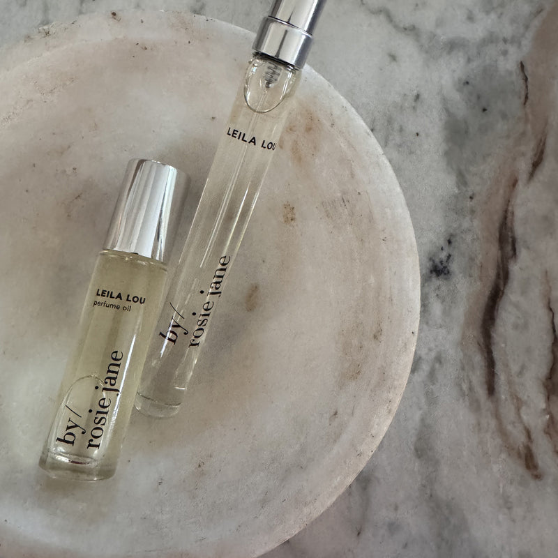 Two glass bottles from the byrosiejane LEILA LOU 2 WAYS TRAVEL SET—one upright, one lying down—rest on a round white dish atop marble, making them the perfect travel perfume companions for life on the go.