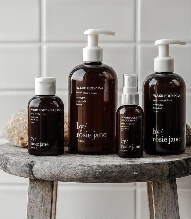 Four amber bottles of By Rosie Jane Wake body products, including body wash, body milk, bath oil, and deodorant, are arranged on a rustic wooden stool with natural sponges in the background.