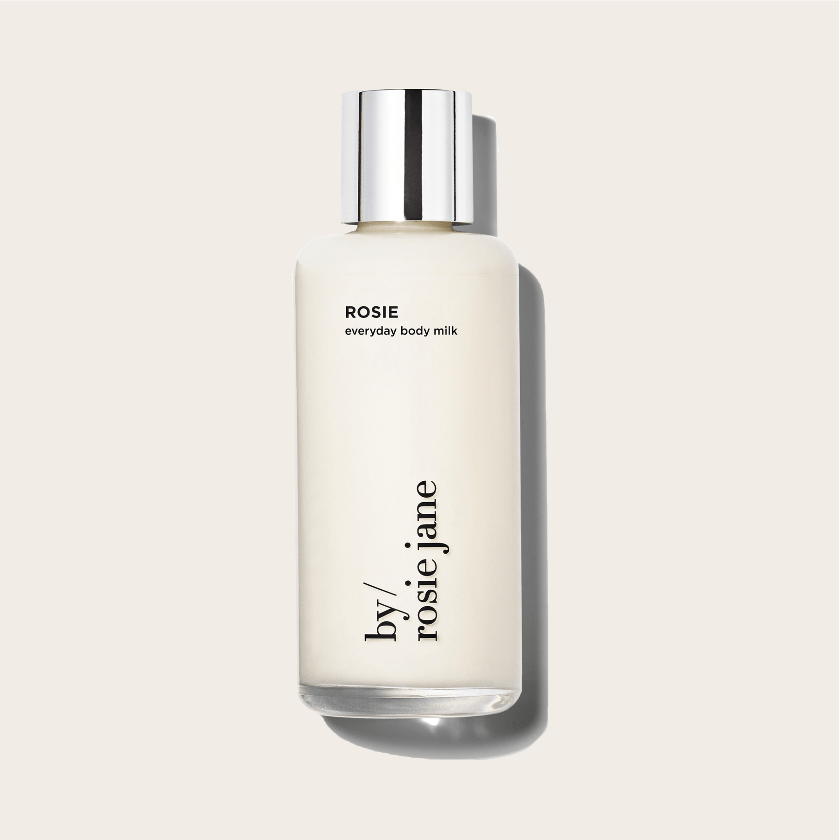 A clear bottle with a silver cap, labeled ROSIE BODY MILK by byrosiejane, contains a creamy, weightless hydrating lotion and stands upright against a light beige background.