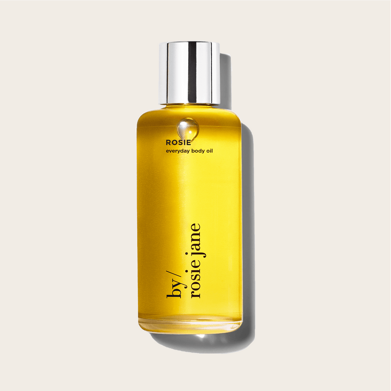A clear glass bottle of byrosiejane ROSIE BODY OIL with a silver cap, filled with golden yellow oil, stands upright on a light beige background.