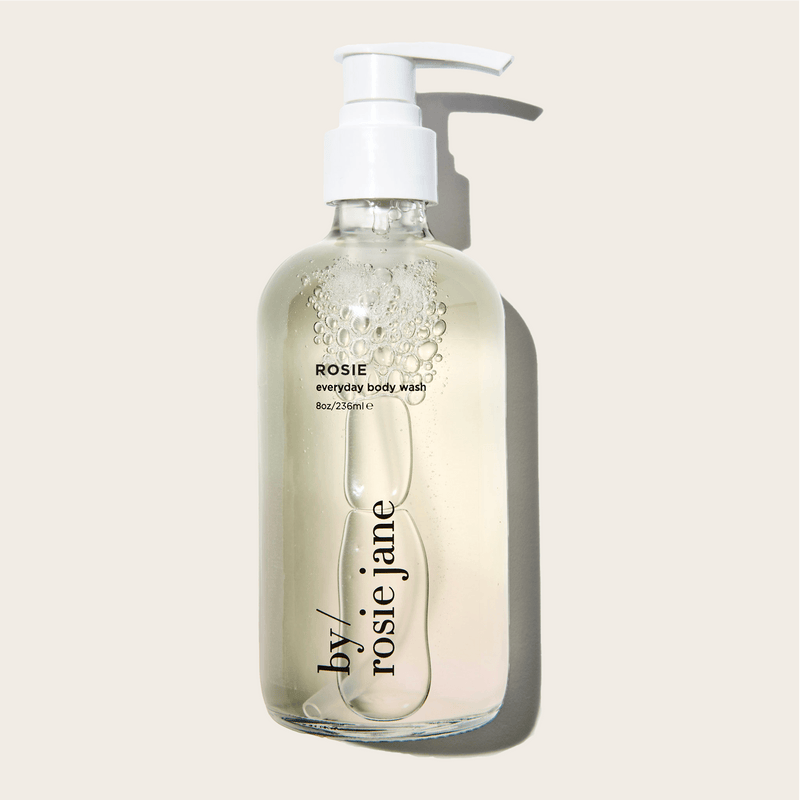 A clear pump bottle labeled byrosiejane contains translucent ROSIE BODY WASH with visible bubbles. The label reads ROSIE BODY WASH 8oz/236ml. The bottle casts a shadow on a light background.