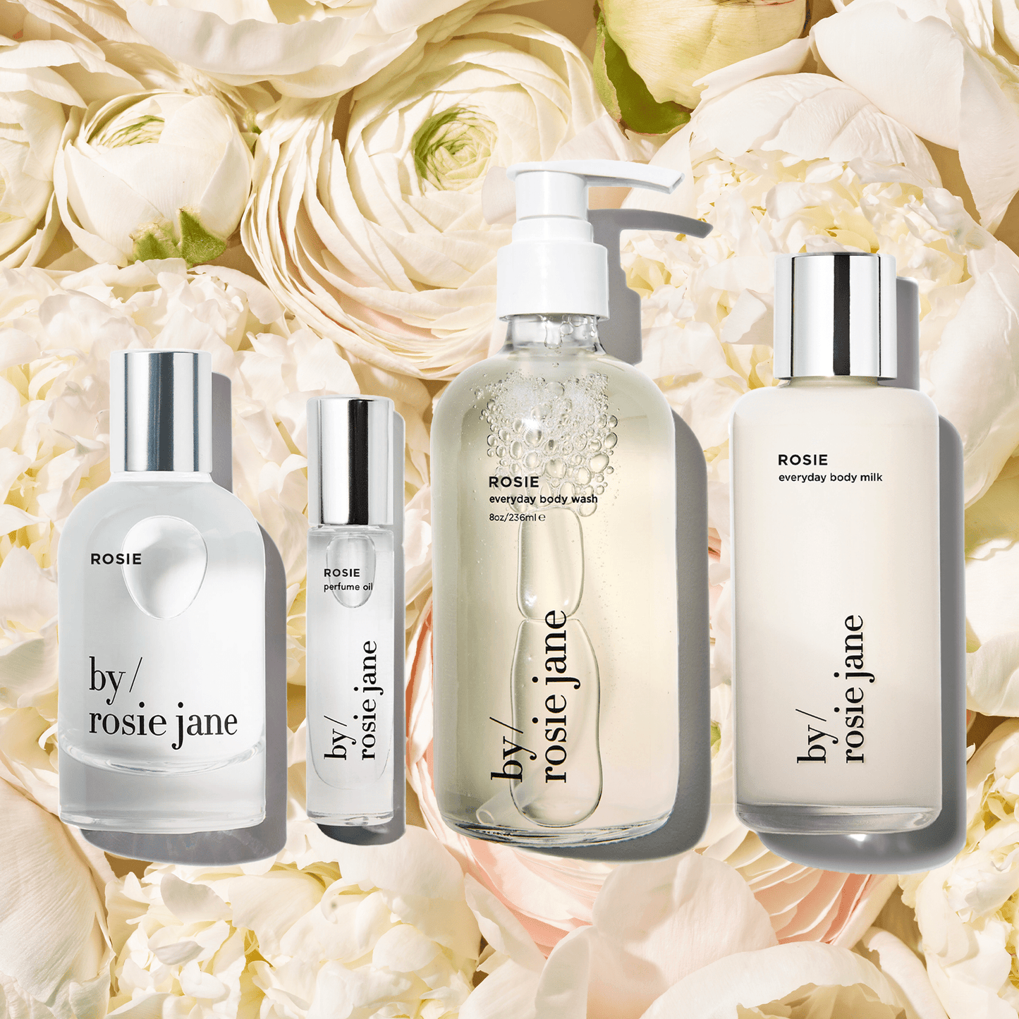 The byrosiejane ROSIE EVERYTHING BUNDLE—two spray bottles, a pump soap bottle, and a body milk—sits before cream and white roses, exuding signature by/rosie jane elegance.