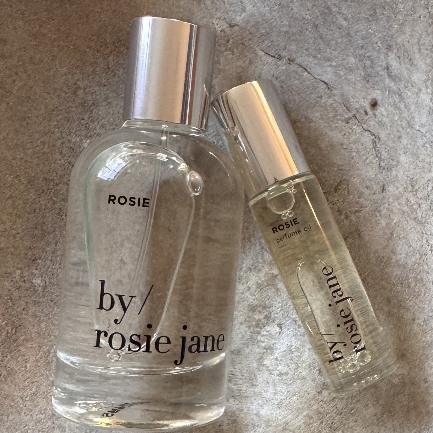 Two clear glass bottles from byrosiejanes LAST ALL DAY SET—one Eau de Parfum and one Perfume Oil—stand side by side on a textured gray surface, delivering long-lasting fragrance in both forms.