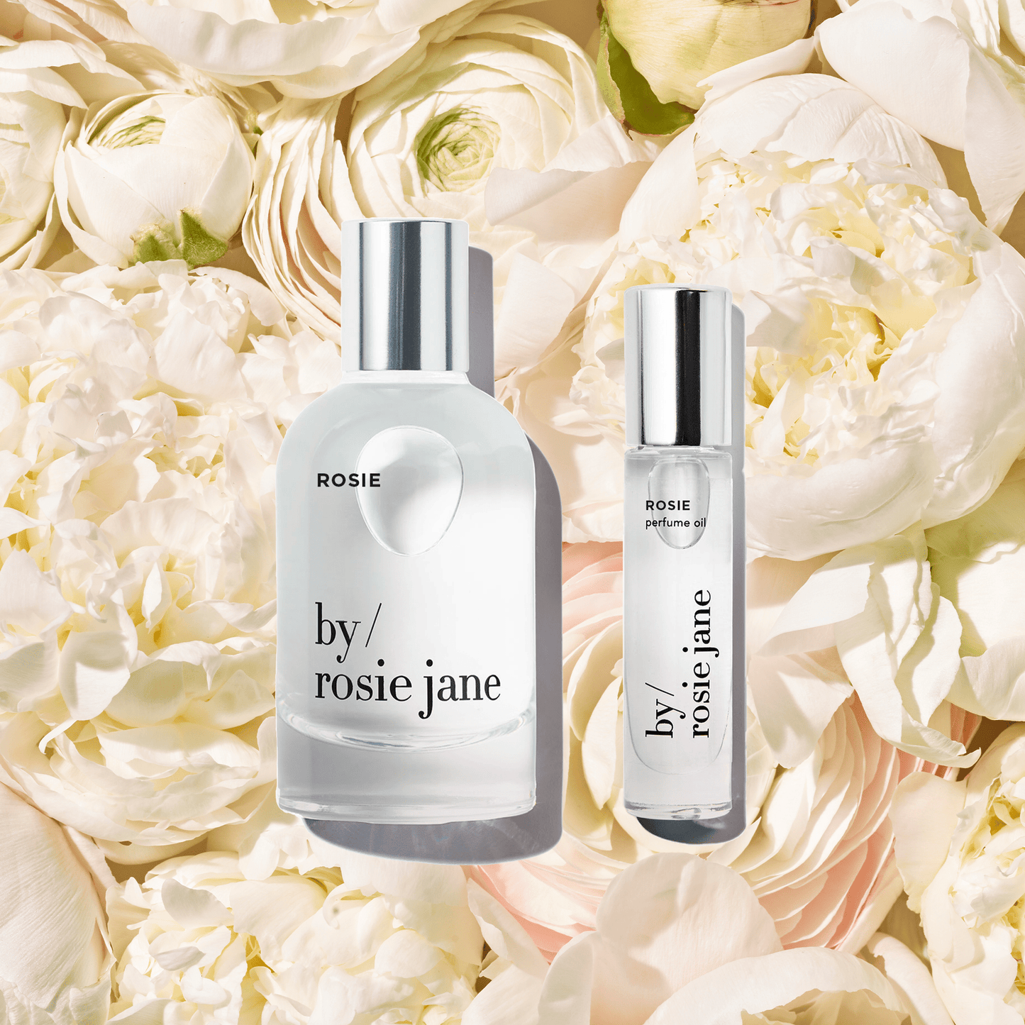 The byrosiejane ROSIE LAST ALL DAY SET features two glass Eau de Parfum bottles, one large and one small, with silver caps and minimal labels, displayed among white and pale pink flowers. This set offers a beautiful, long-lasting fragrance.