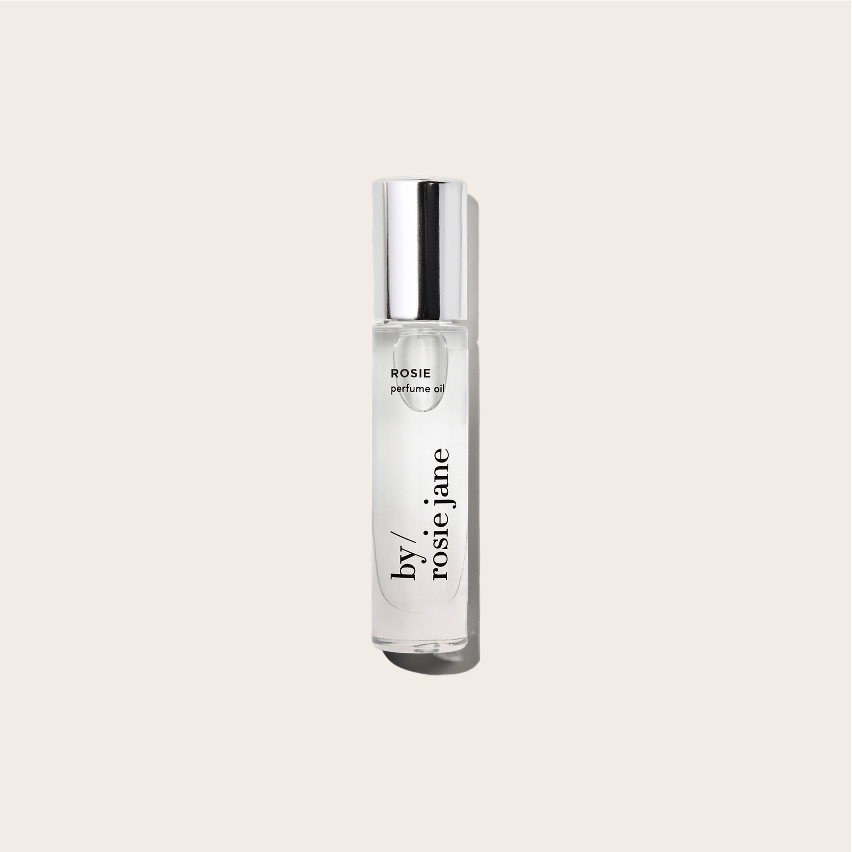 A clear glass bottle of byrosiejane ROSIE PERFUME OIL with a silver cap stands upright against a light beige background. The fragrance features delicate rose oil combined with subtle nude musk notes.