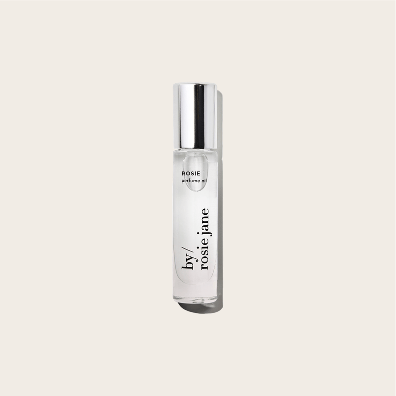 A clear glass bottle of byrosiejane ROSIE PERFUME OIL with a silver cap stands upright against a light beige background. The fragrance features delicate rose oil combined with subtle nude musk notes.