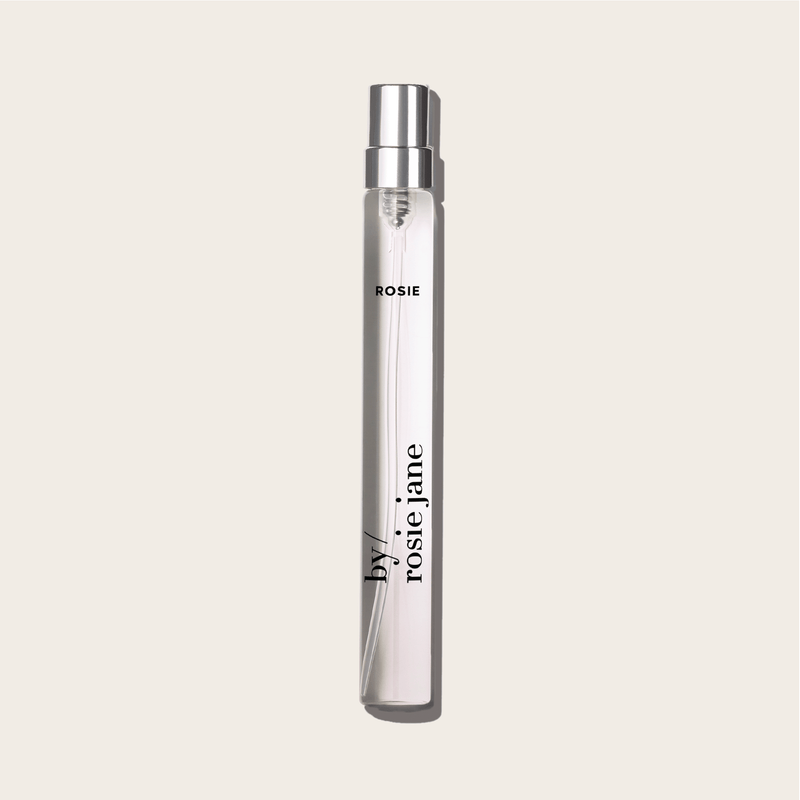 A clear glass perfume spray bottle labeled byrosiejane with ROSIE TRAVEL SPRAY near the top, featuring notes of nude musk, set against a light beige background.