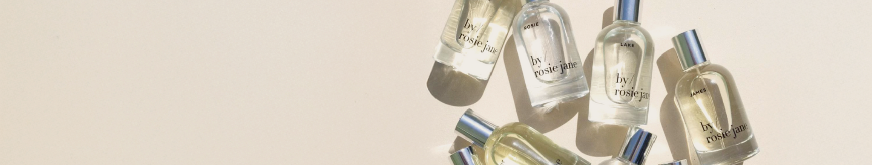 Several clear glass perfume bottles with shiny silver caps are arranged on a light, neutral surface, casting soft shadows. The bottles are labeled by rosie jane.