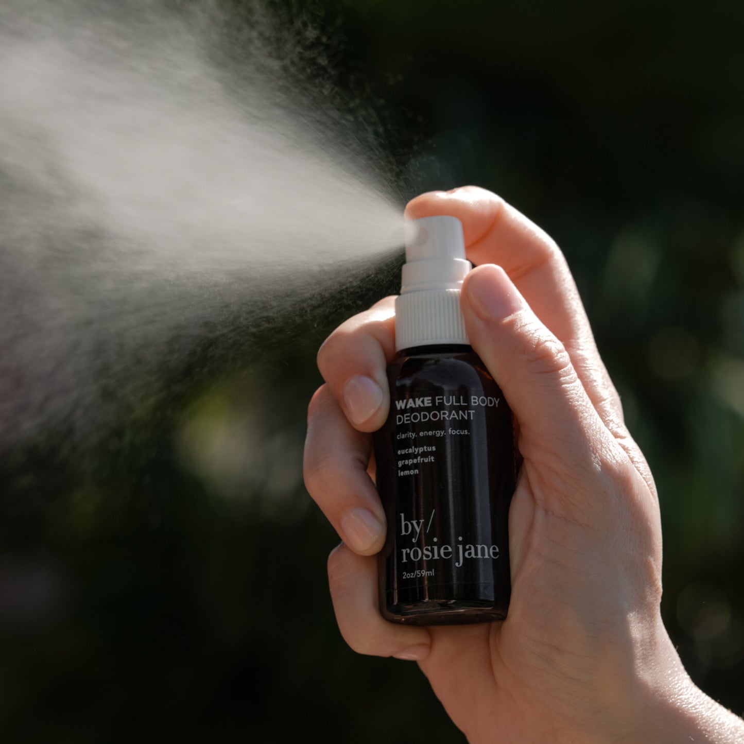 A hand sprays the black byrosiejane WAKE FULL BODY DEODORANT bottle, releasing a visible mist from its white pump against a dark background, highlighting this natural deodorant for sensitive skin.