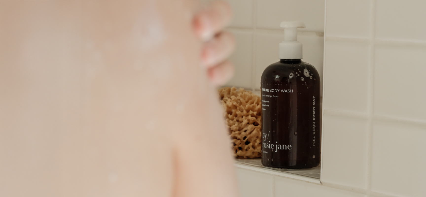 A close-up of a person showering, with a bottle of body wash and a natural sponge on a tiled shelf in the background. The bottle is dark with a white pump and labeled body wash.