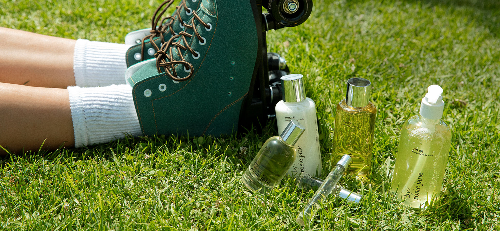 A person wearing white socks and green roller skates sits on grass beside various bottles of skincare or beauty products.