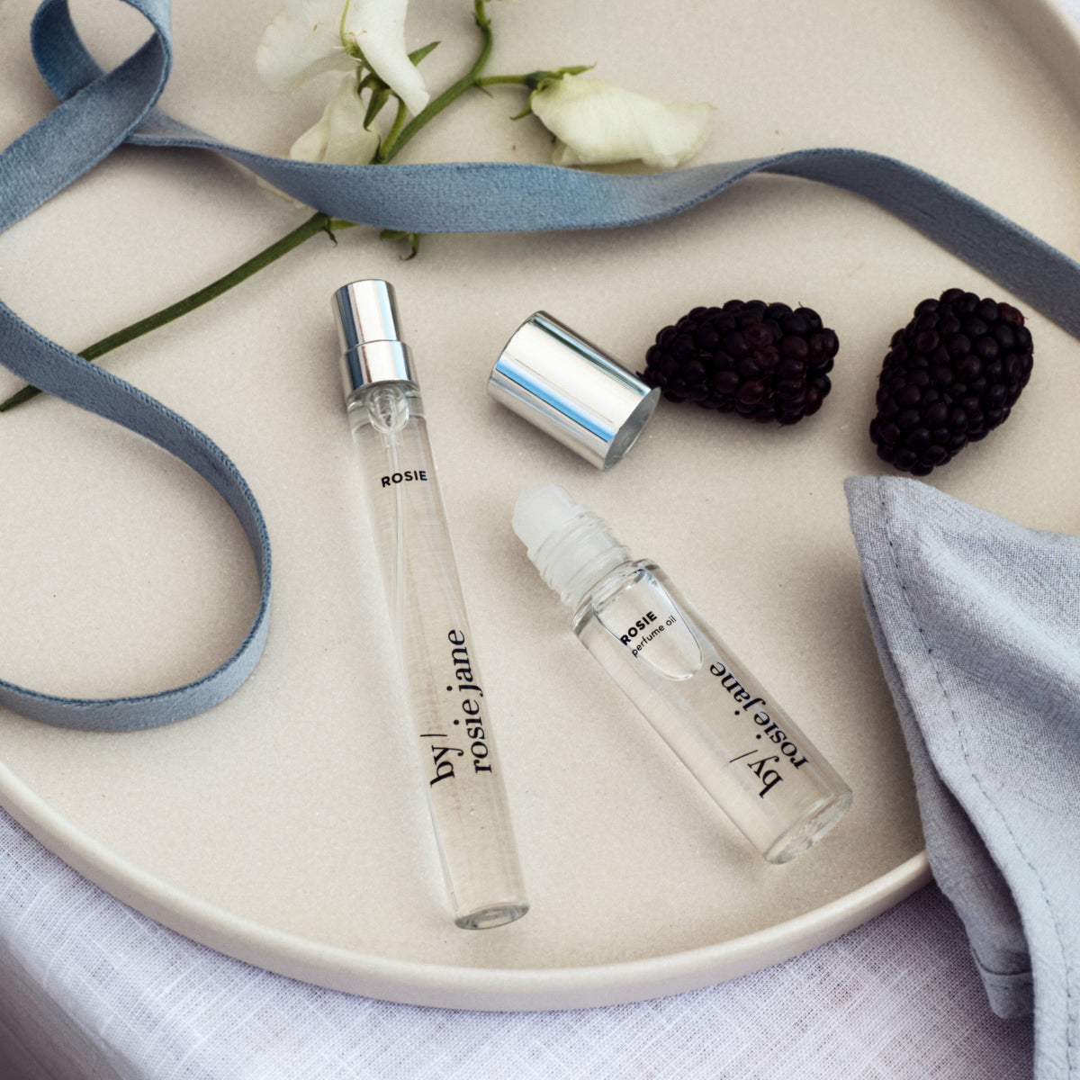 Two clear glass rollerball perfume sample vials with silver caps from 'by rosie jane' are arranged on a beige ceramic plate with a blue ribbon, white flowers, blackberries, and a folded light blue cloth.