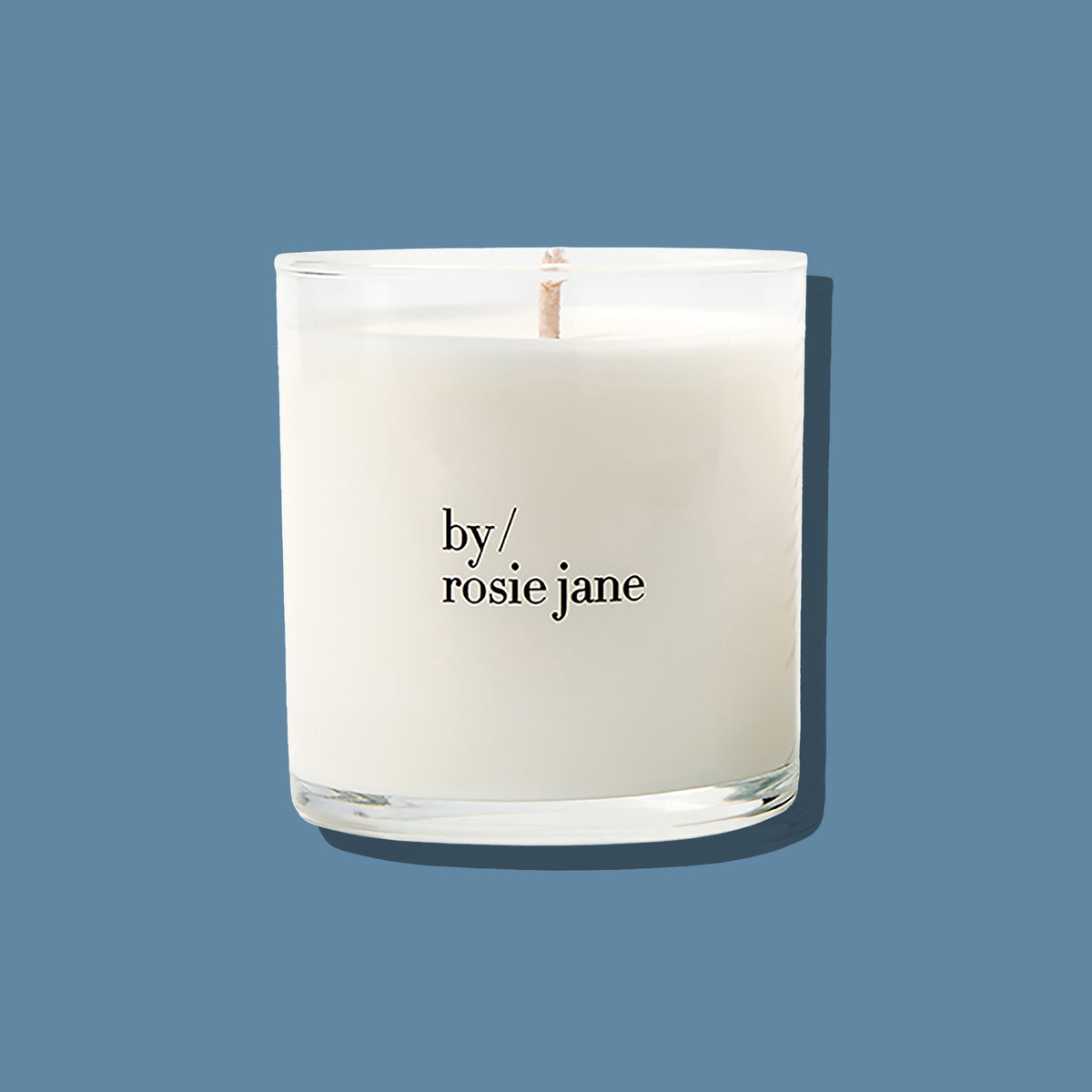 A white scented candle in a clear glass jar with a single wick, printed with the brand name 'by/ rosie jane' in black text, set against a solid slate blue background.