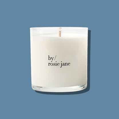A white scented candle in a clear glass jar with a single wick, printed with the brand name 'by/ rosie jane' in black text, set against a solid slate blue background.