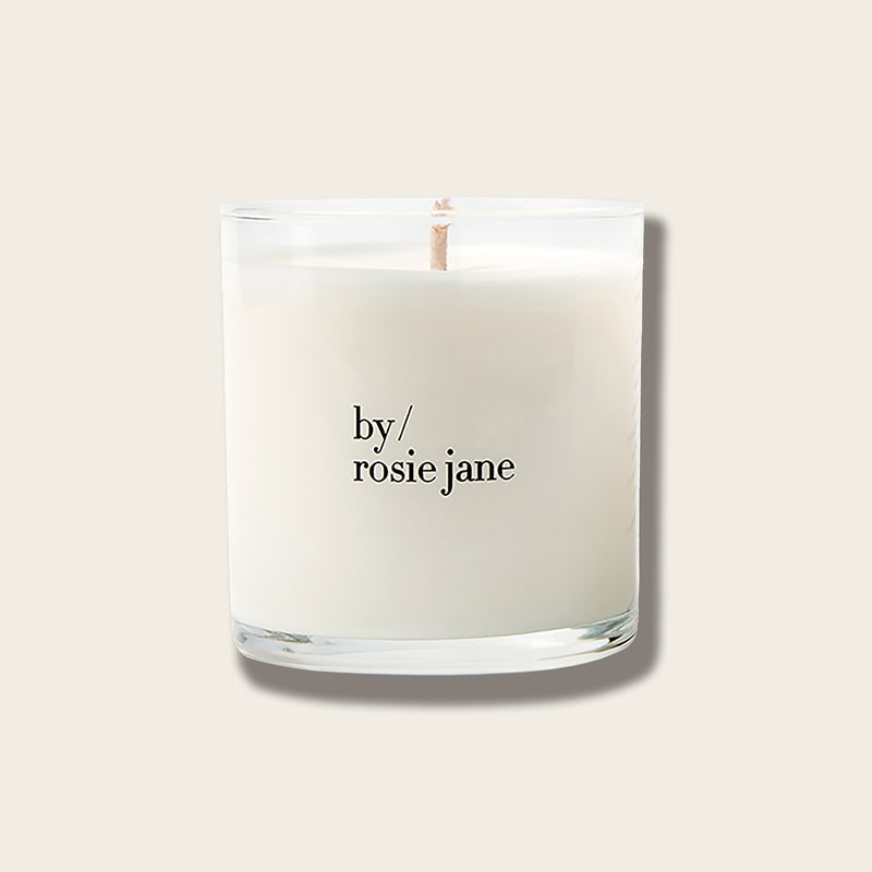 A white scented candle in a clear glass jar with a single wick, featuring the text 'by/ rosie jane' printed in black on the front, set against a solid light beige background.