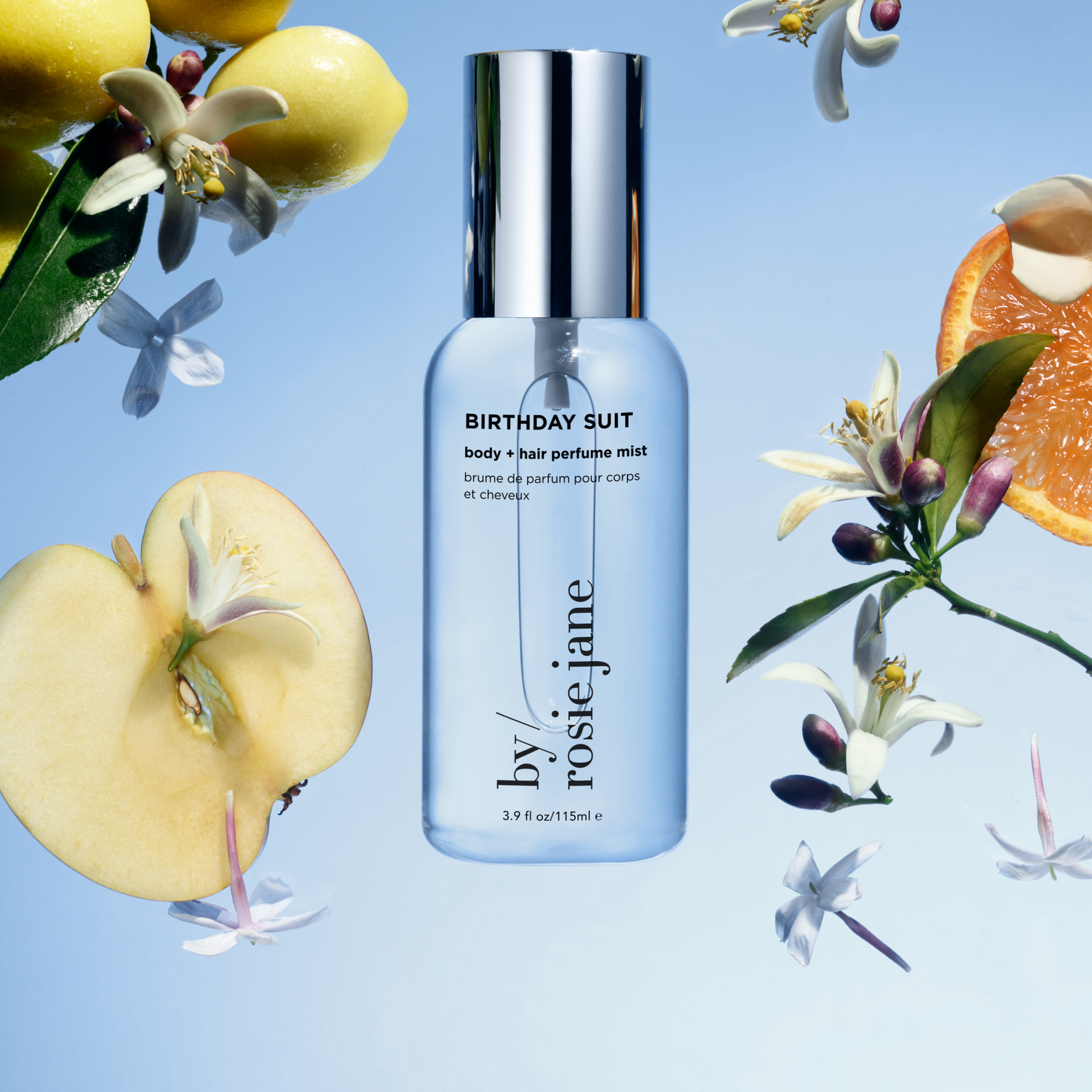 A clear bottle of Birthday Suit body and hair perfume mist by Rosie Jane is surrounded by apple slices, orange slices, white flowers, and green leaves on a blue background.