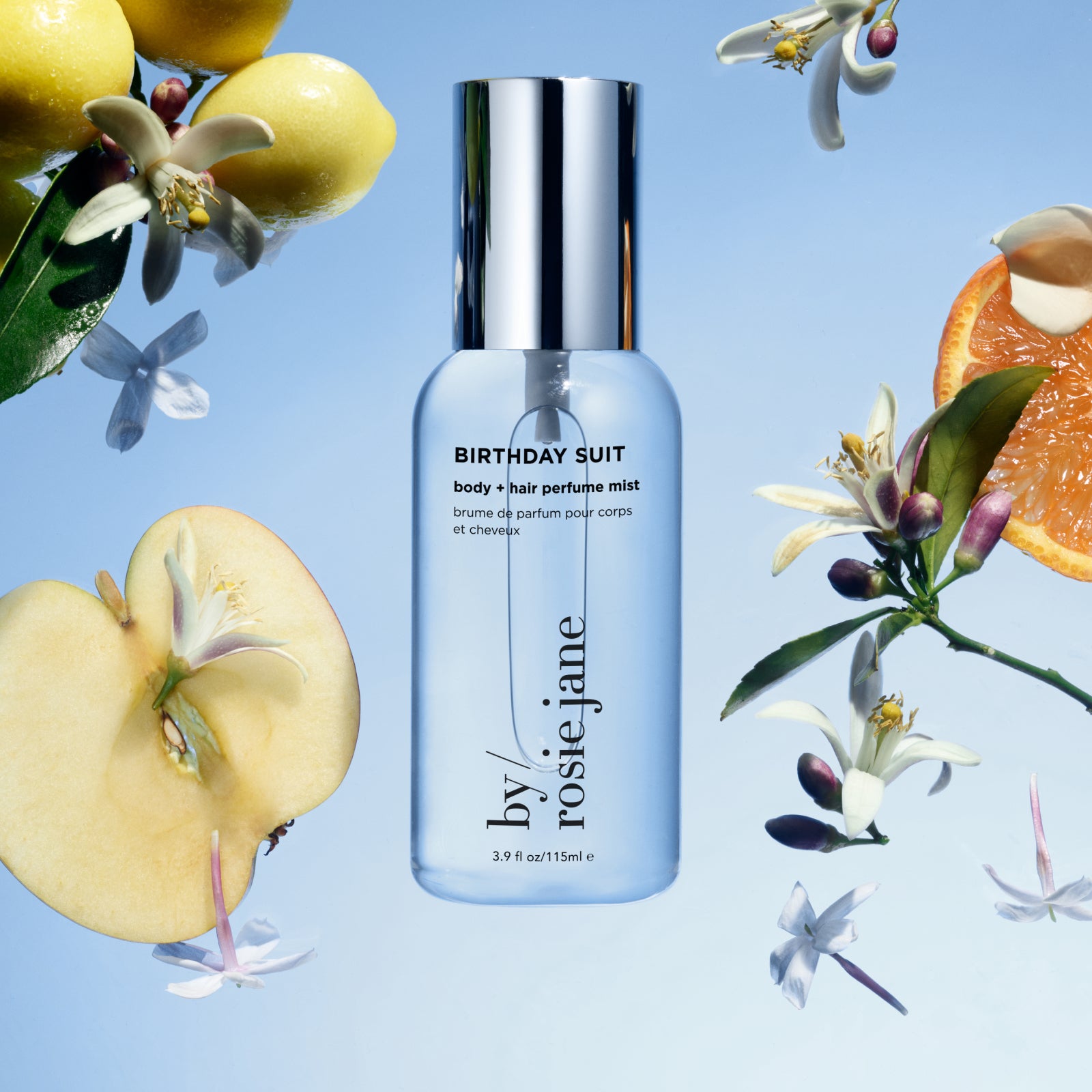 A clear bottle of Birthday Suit body and hair perfume mist by Rosie Jane is surrounded by apple slices, orange slices, white blossoms, and green leaves, all set against a light blue background.
