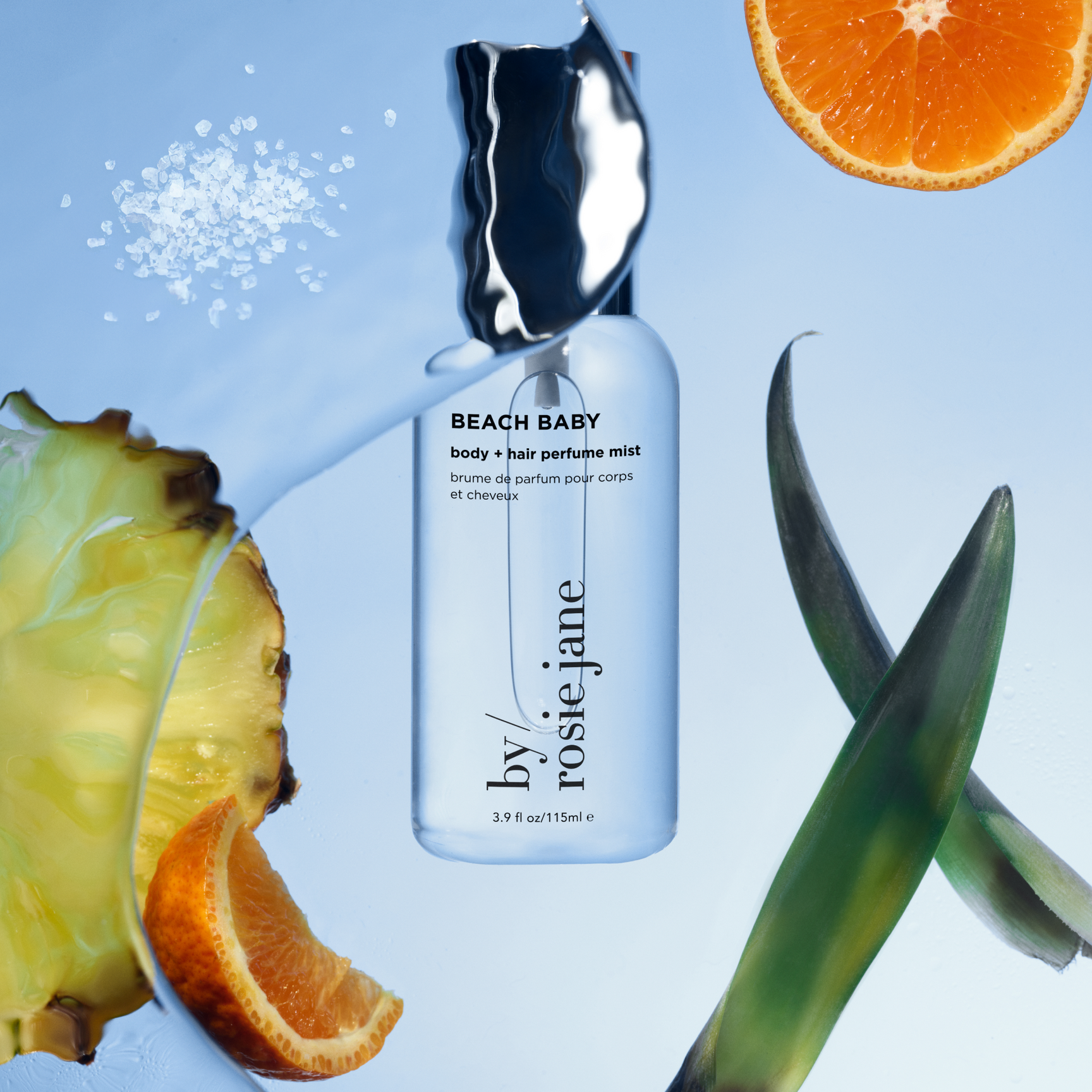 A bottle of Beach Baby body + hair perfume mist by Rosie Jane is surrounded by orange slices, pineapple, green leaves, and sea salt against a blue background.