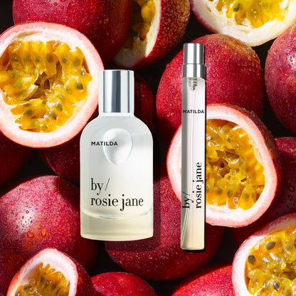 Matilda by Rosie Jane perfume bottle and matching travel spray arranged on a background of fresh red passion fruit halves.