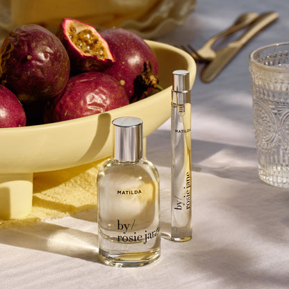 Two perfume bottles labeled 'by rosie jane' on a table with fruits in the background