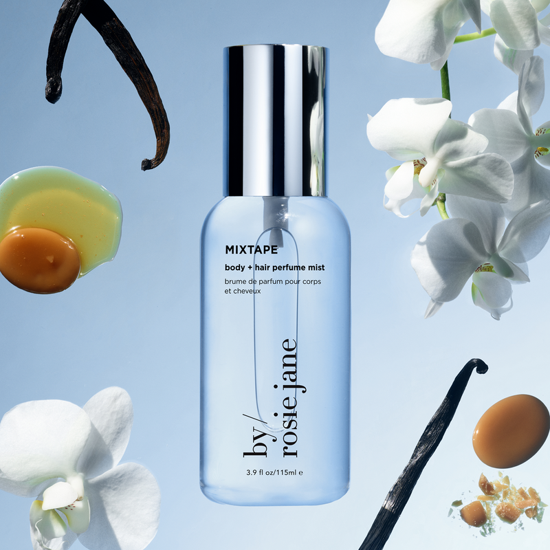 A bottle of ROSIE JANE MIXTAPE body + hair perfume mist centered on a light blue background, surrounded by floating vanilla beans, white orchid flowers, and vanilla seeds. The bottle is clear with a silver cap and black text.