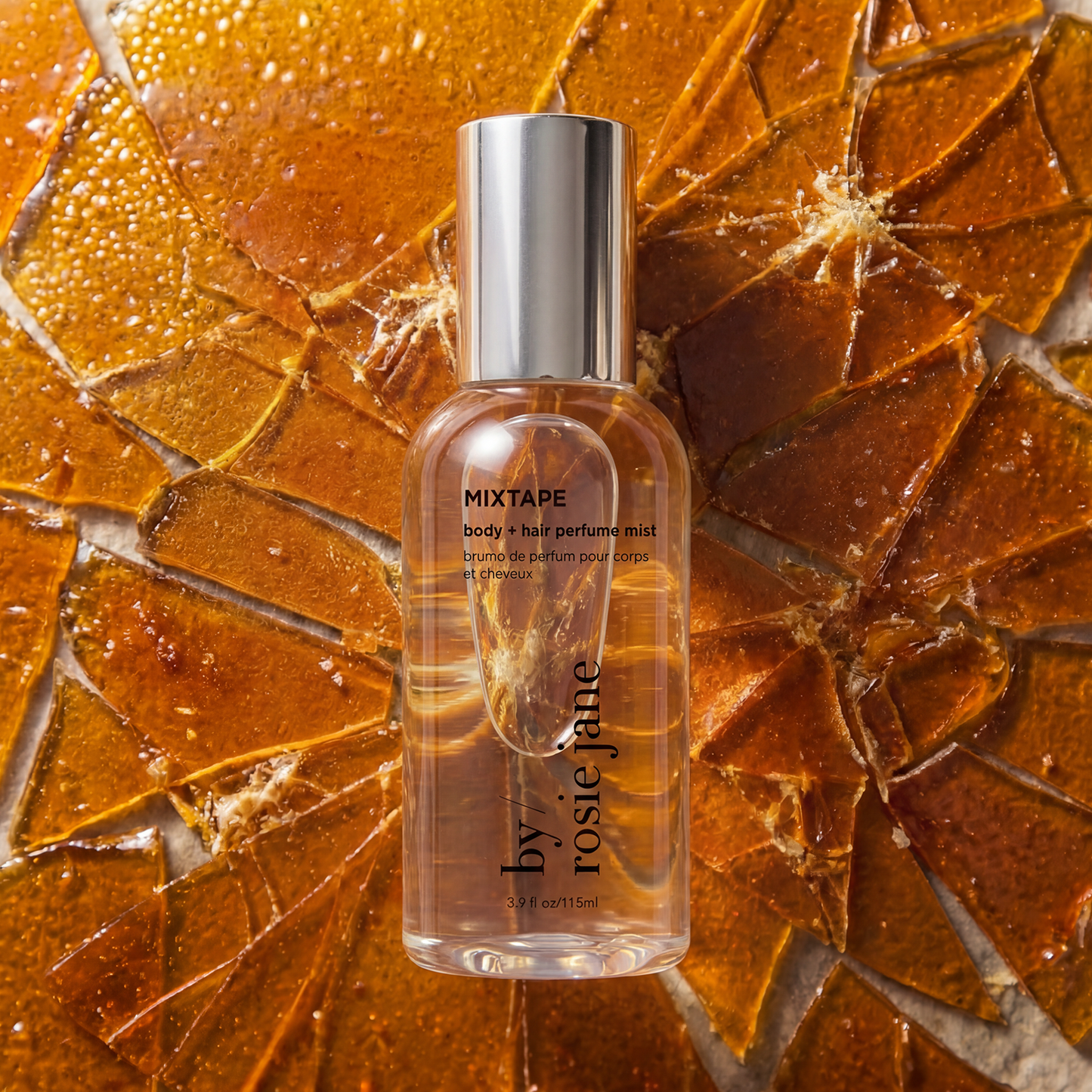 A clear bottle of by rosie jane Mixtape body and hair perfume mist with a silver cap, set against a textured background of golden-brown cracked butterscotch.