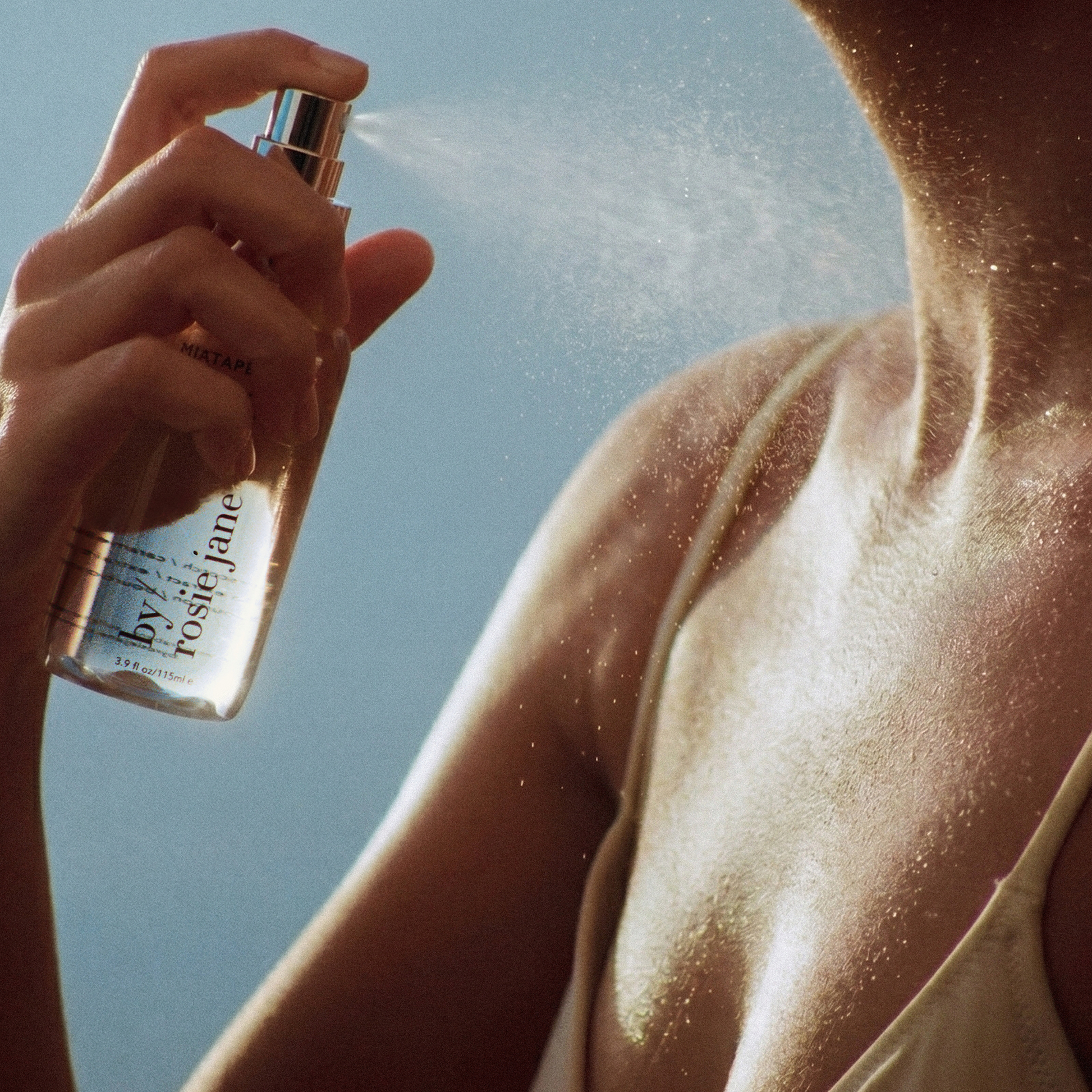 A close-up lifestyle shot of a person spraying a clear bottle of Rosie Jane sunscreen onto their skin, with fine droplets visible against a bright blue sky.