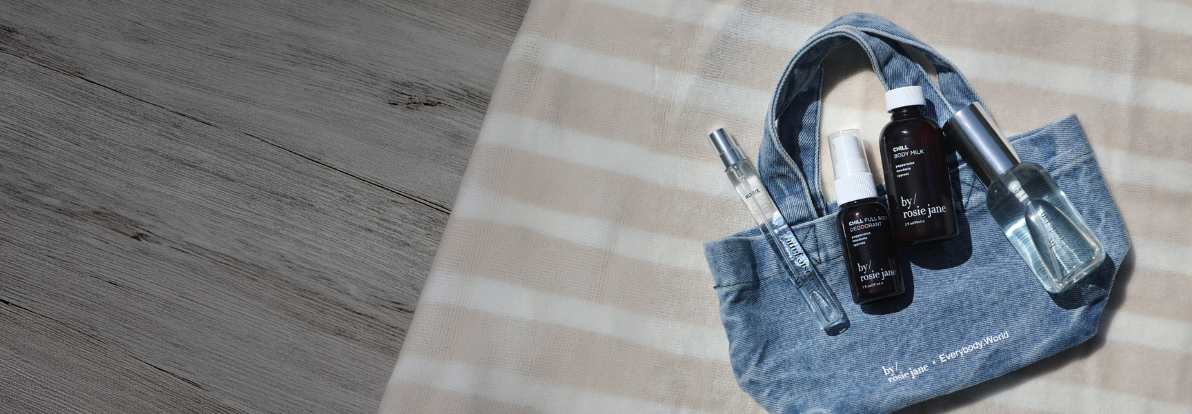 A blue denim tote bag laid on a beige-and-white checkered blanket over a gray wood floor, holding two black skincare bottles and a clear glass tube with a silver cap.