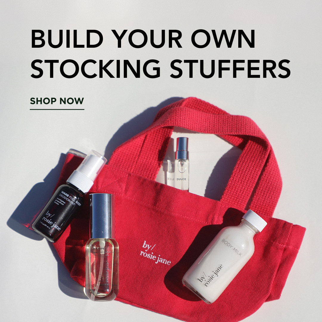 A red canvas stocking stuffer bag with white 'by rosie jane' logos, styled with several skincare and fragrance products on a light gray background. Large text at the top reads 'BUILD YOUR OWN STOCKING STUFFERS' with 'SHOP NOW' below.