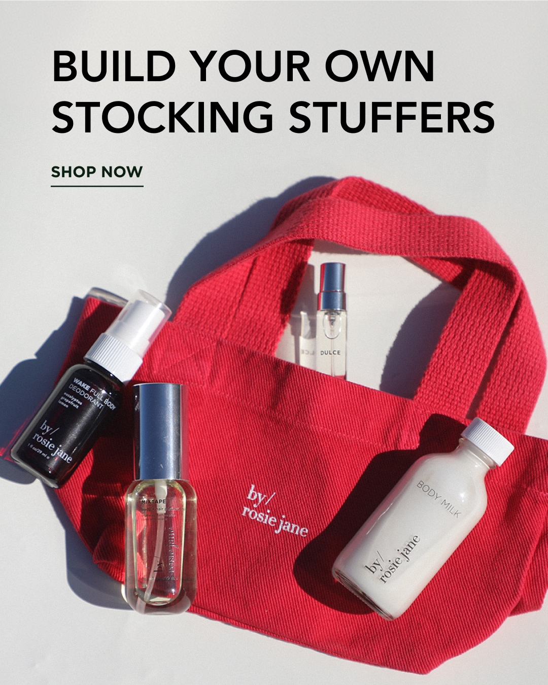 A red canvas tote bag with the words 'by / rose jane' printed on the front pocket, styled with several skincare and body products from the same brand. Large text at the top reads 'BUILD YOUR OWN STOCKING STUFFERS' with 'SHOP NOW' below, on a clean white background.
