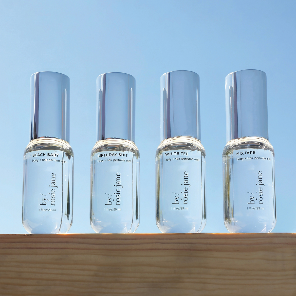 Four small clear perfume bottles with reflective silver caps are lined up on a wooden surface against a clear blue sky. The bottles are labeled by rose jane and feature scents: Beach Baby, Birthday Suit, White Tee, and Mixtape.