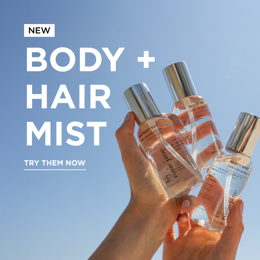 A hand holds up three bottles of body and hair mist against a clear blue sky, with text that reads NEW Body + Hair Mist. Try them now. Each bottle has a silver cap and clear liquid inside.