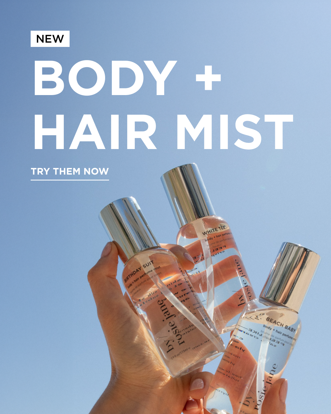 A hand holds up three clear bottles of body and hair mist with silver caps against a blue sky. Large white text reads NEW Body + Hair Mist. Try them now.