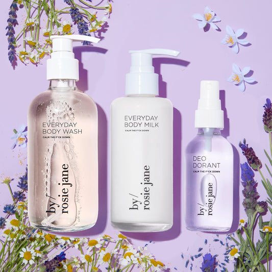 Calm The F*ck Down Complete Body Collection, featuring bottles of deodorant, body milk, and body wash, surrounded by flowers, designed for comprehensive skin care.