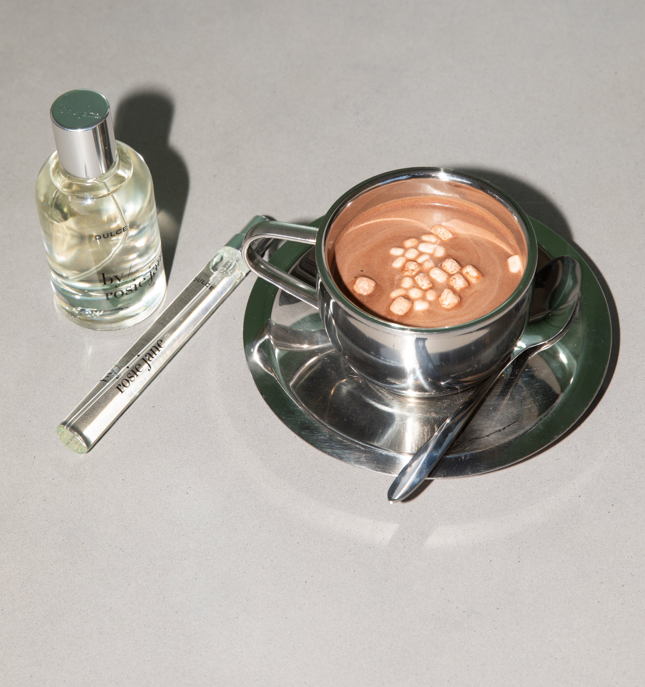 Silver cup and saucer with a two brj perfume bottles on a light gray background
