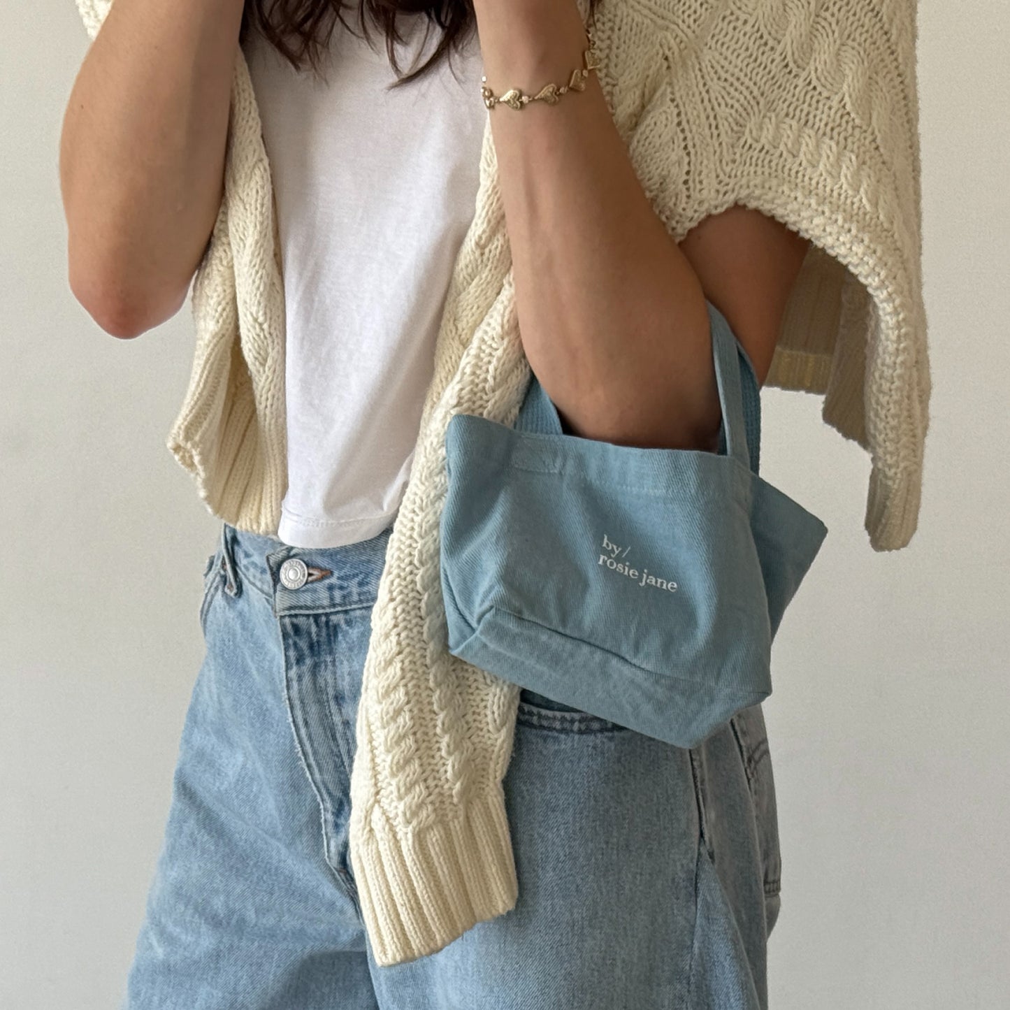 Person wearing a beige cardigan, white shirt, blue jeans, and holding a blue tote with 'by rose jane' branding.