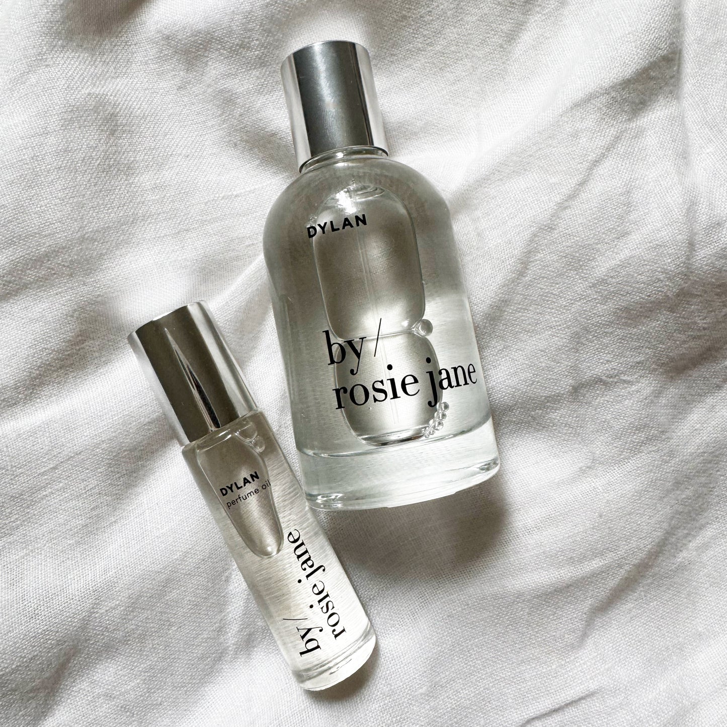 Two clear glass perfume bottles with silver caps, labeled DYLAN by byrosiejane, rest on white fabric. The DYLAN LAST ALL DAY SET features a full-size Eau de Parfum and a travel spray for enjoying the long-lasting scent anywhere.