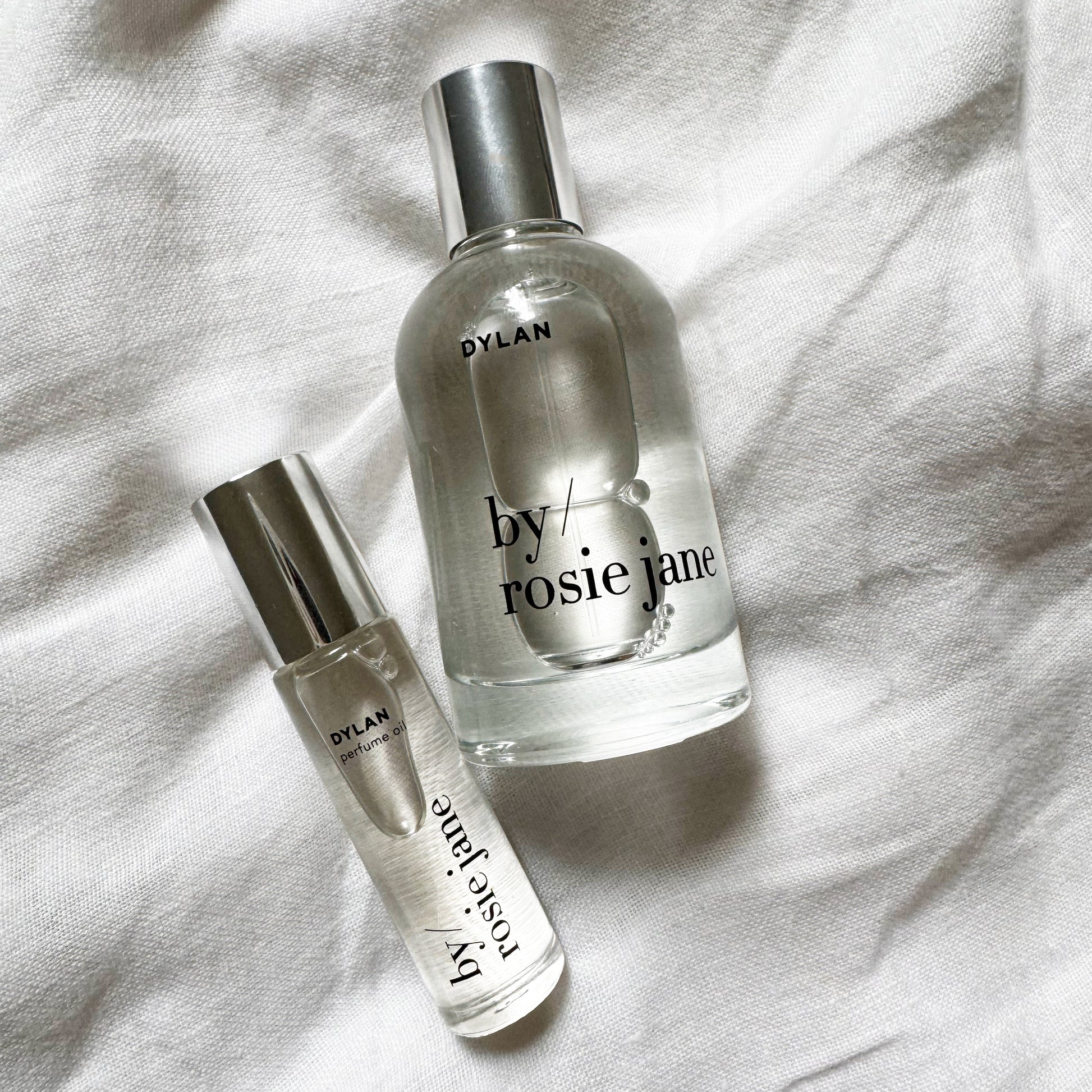 Two clear glass perfume bottles with silver caps, labeled DYLAN by byrosiejane, rest on white fabric. The DYLAN LAST ALL DAY SET features a full-size Eau de Parfum and a travel spray for enjoying the long-lasting scent anywhere.