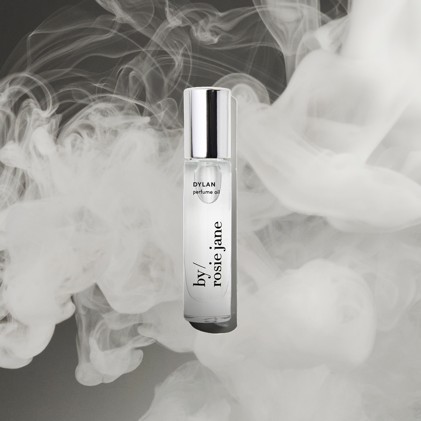 A clear glass DYLAN PERFUME OIL bottle by byrosiejane with a silver cap, centered on a smoky dark gray background with swirling white mist—evoking the allure of white musk and whispers of sexual freedom.